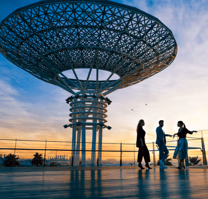 Three friends walking along a boardwalk at Bluewaters Island during sunset, with the island’s iconic steel canopy structure towering above.