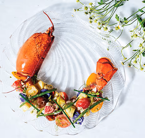 Elegant seafood dish featuring lobster claw and tail served with colorful heirloom tomatoes, vegetables, and fresh herbs on a decorative glass plate.