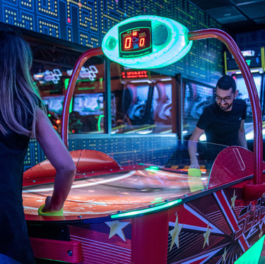 Two people enjoying a game of air hockey in a neon-lit arcade with Pac-Man-themed walls at Bluewaters Island.