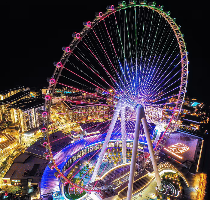 Aerial night view of the Ain Dubai observation wheel illuminated with vibrant rainbow lights, surrounded by the glowing cityscape of Bluewaters Island.