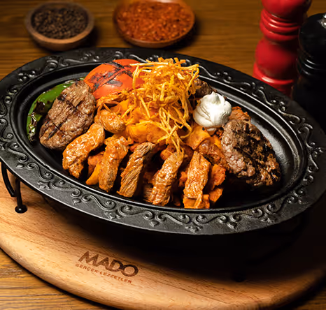 Hot cast iron plate filled with assorted grilled meats, vegetables, and crispy potatoes, served on a wooden board at MADO restaurant in Bluewaters.