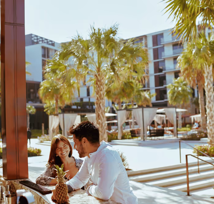 Smiling couple sitting at an outdoor bar with a tropical drink, surrounded by palm trees and luxury resort cabanas.