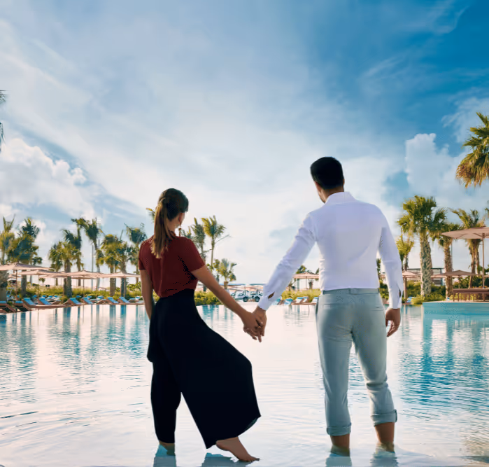 Couple holding hands while standing ankle-deep in a luxury resort pool, surrounded by palm trees and sun loungers.