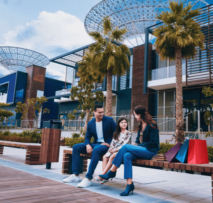 Man, woman, and young girl sitting on a bench with shopping bags in an outdoor plaza under Bluewaters’ signature steel canopies.