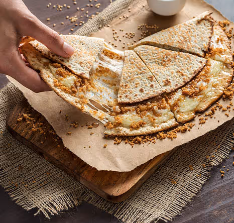 Close-up of a hand pulling a slice of warm flatbread filled with melted cheese and sweet crumble topping, served on brown paper over a wooden board.