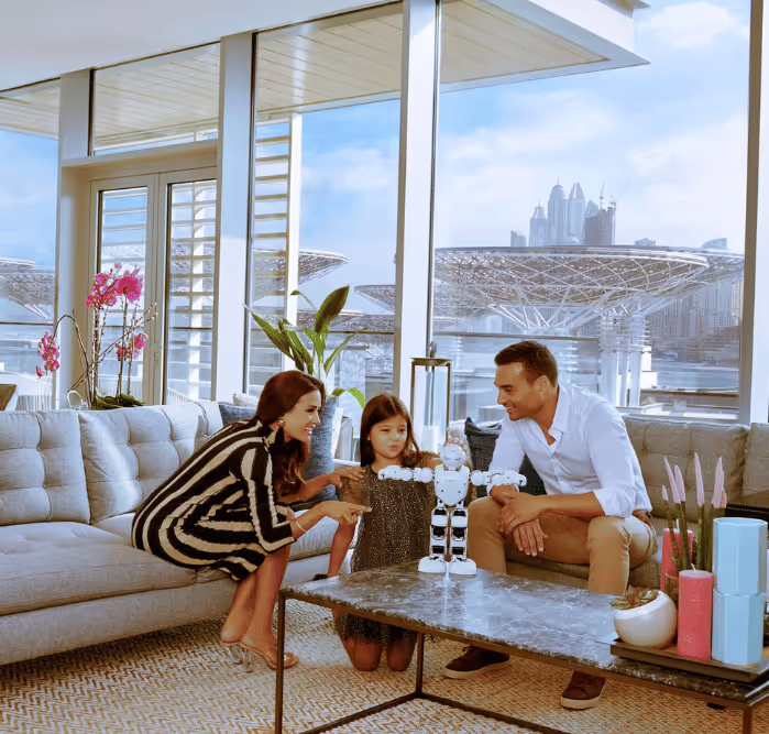Smiling couple and young girl playing with a toy robot in a bright living room with floor-to-ceiling windows overlooking Bluewaters Island and Dubai Marina skyline.