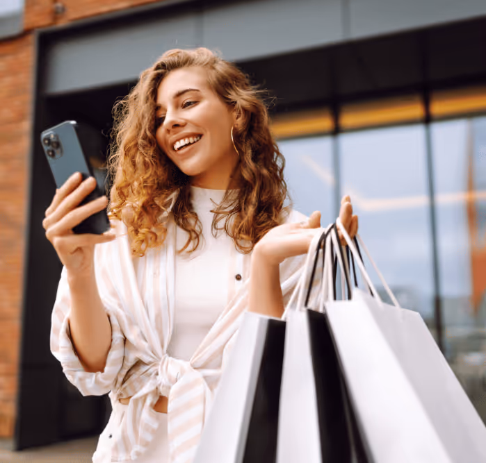 Young woman holding shopping bags and looking at her phone with a smile outside Bluewaters shopping area.
