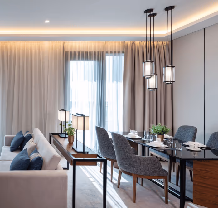Stylish open-plan dining and living space with a glass-top table, upholstered chairs, pendant lights, and a cream sofa, accented by soft curtains and natural light.