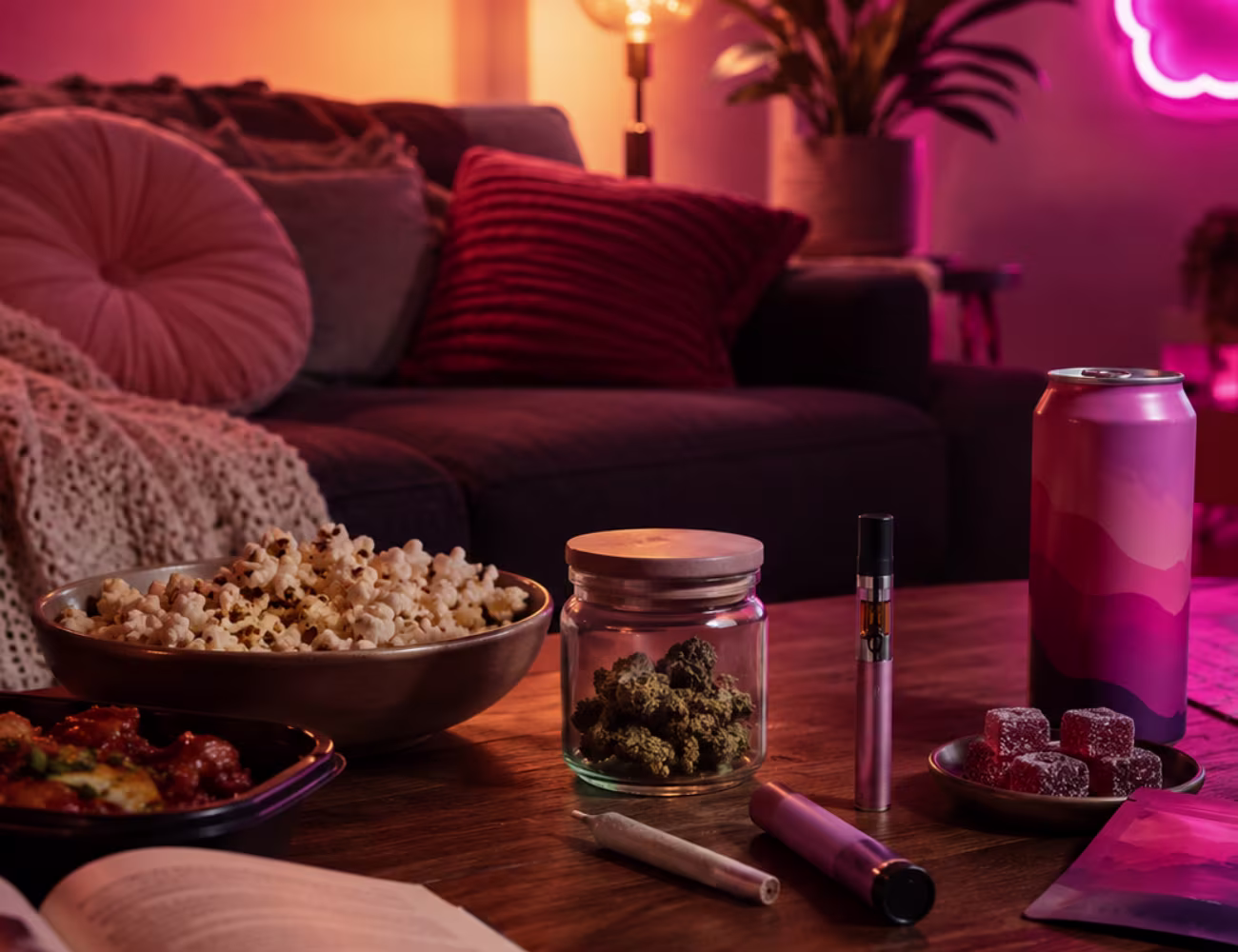 Bright, cozy living room with a soft blanket, lamp, movie snacks, a mug, books, and neatly displayed flower, pre-roll, vape, edible, and beverage packages on a coffee table.
