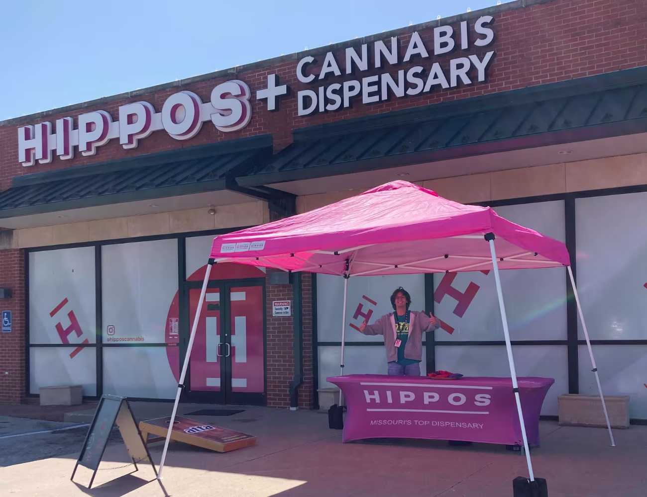 Hippos Cannabis Dispensary storefront with a staff member standing beneath a bright pink branded canopy beside a cornhole game on a sunny day.