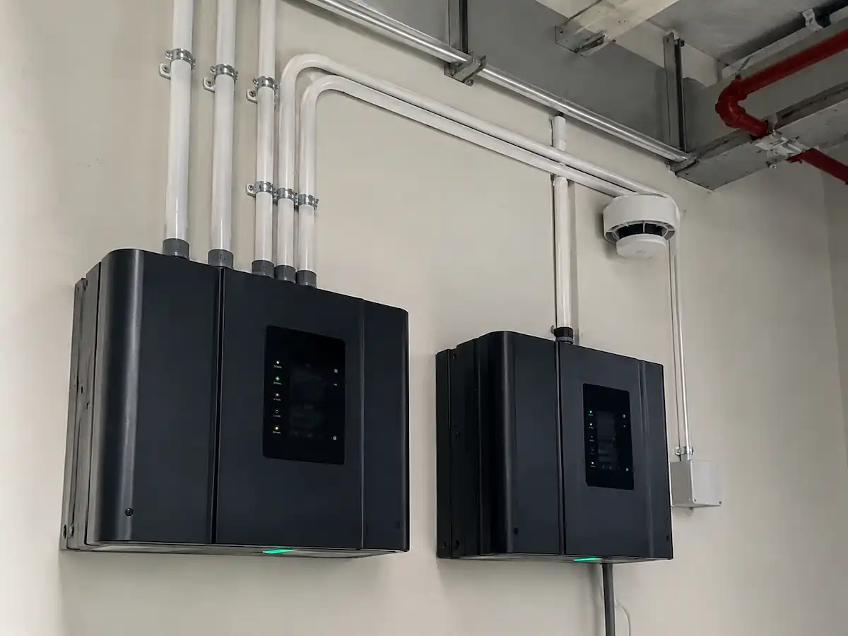 Two black electronic control panels mounted on a wall with connected white pipes and a round sensor above.