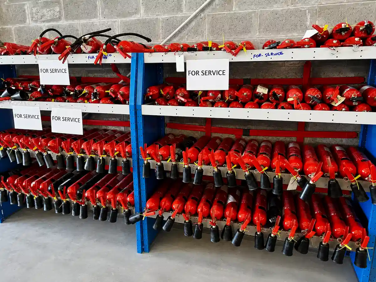 Shelves filled with red fire extinguishers labeled with multiple 'FOR SERVICE' signs in a storage area.