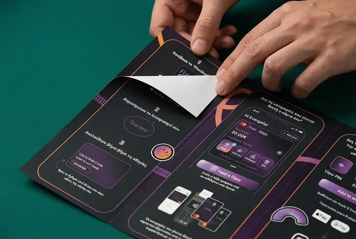 Χέρια που ξεκολλούν το προστατευτικό χαρτί από μια κάρτα πληρωμής με εμφανή ψηφιακή εφαρμογή σε μαύρο φόντο.