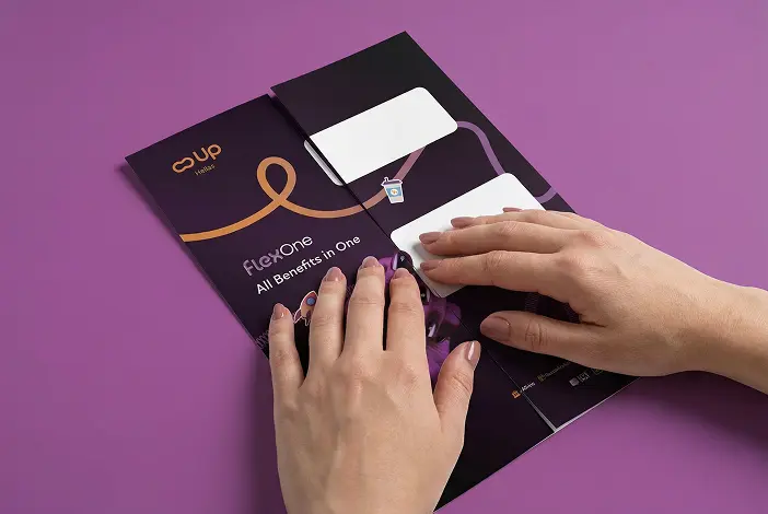 Χέρια διπλώνουν ένα φυλλάδιο με μαύρο και μοβ φόντο που έχει τίτλο FlexOne και φράση All Benefits in One.