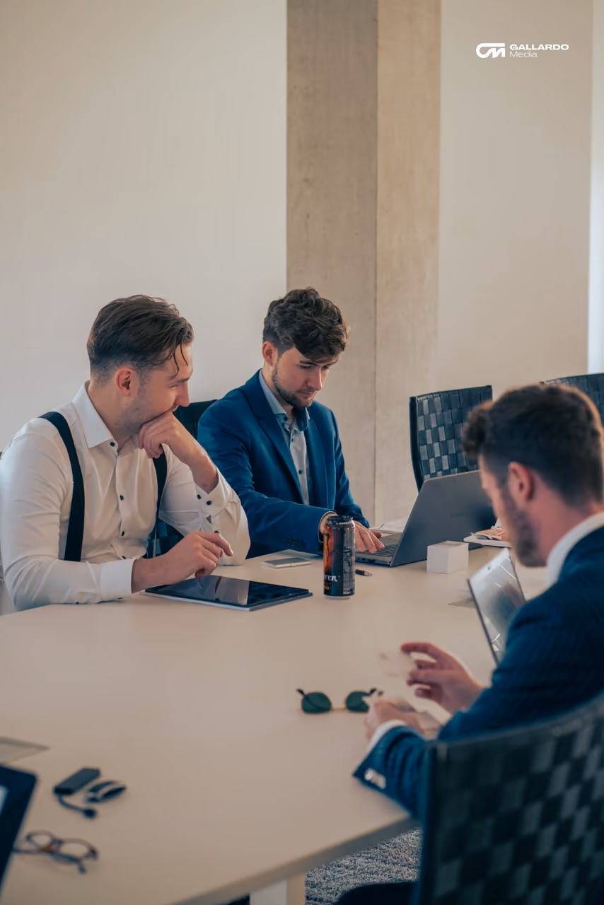 Drei Männer in formeller Kleidung arbeiten an Laptops und einem Tablet um einen Konferenztisch.
