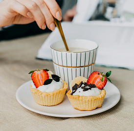 Two mini tartlets topped with whipped cream, fresh strawberries, and chocolate shavings on a white plate, served alongside a striped cup of coffee being stirred with a golden spoon.