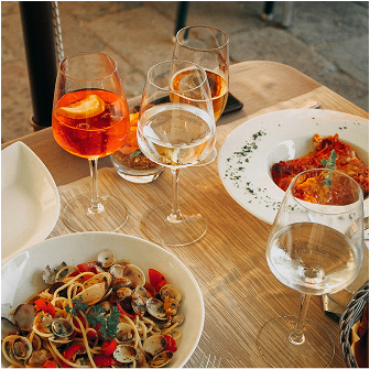 A table set with plates of seafood pasta and ravioli, accompanied by glasses of sparkling Aperol Spritz and white wine, at an outdoor dining area.