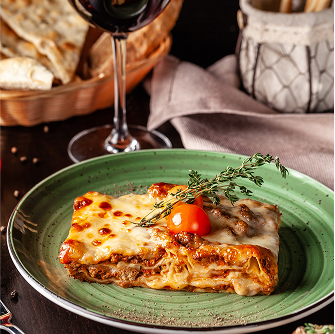 A slice of baked lasagna topped with cherry tomatoes and herbs, served on a green plate, accompanied by a glass of red wine and a basket of flatbread in the background.