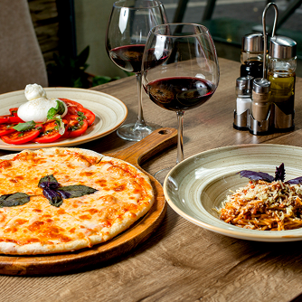 A wooden table set with a Margherita pizza, a plate of spaghetti topped with cheese and basil, a tomato and mozzarella Caprese salad, and two glasses of red wine in a cozy restaurant setting.
