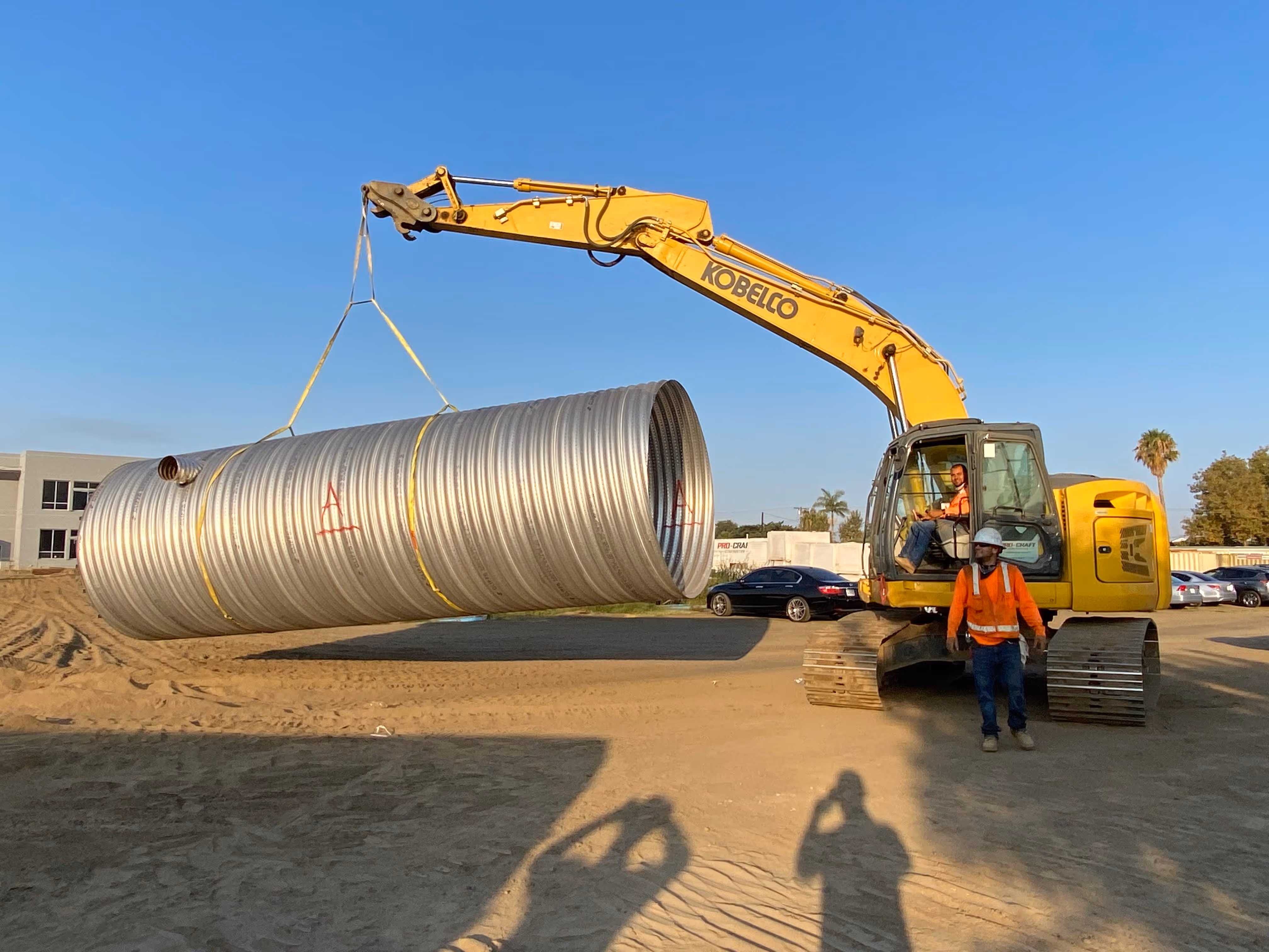 Excavator crane lifting large corrugated metal culvert pipe at construction site.