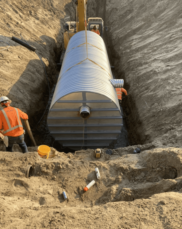 Construction workers lowering a large corrugated metal tank into a trench for underground utility installation.