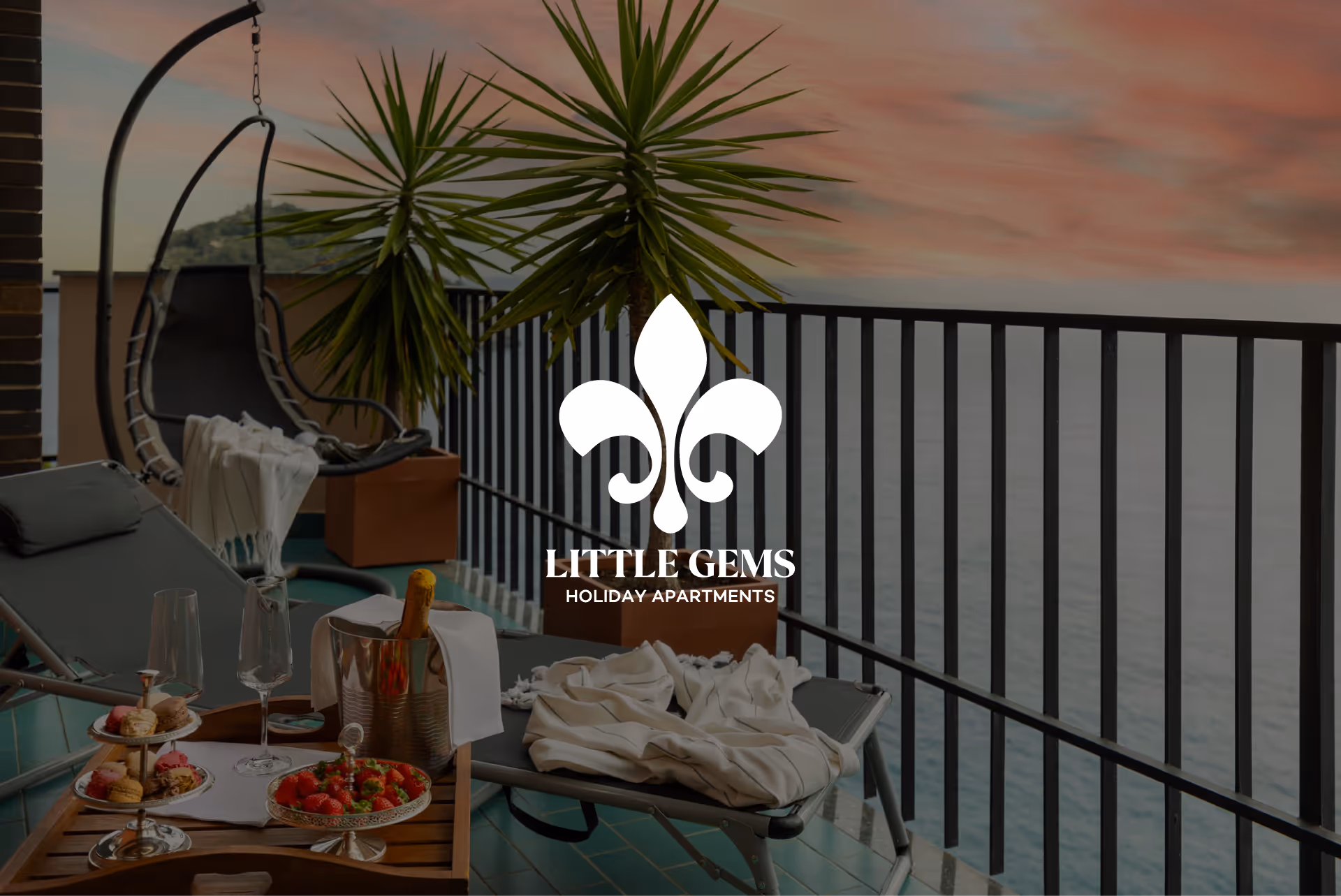 Balcone con tavolo, bevande, frutta, piante e logo Little Gems Holiday Apartments