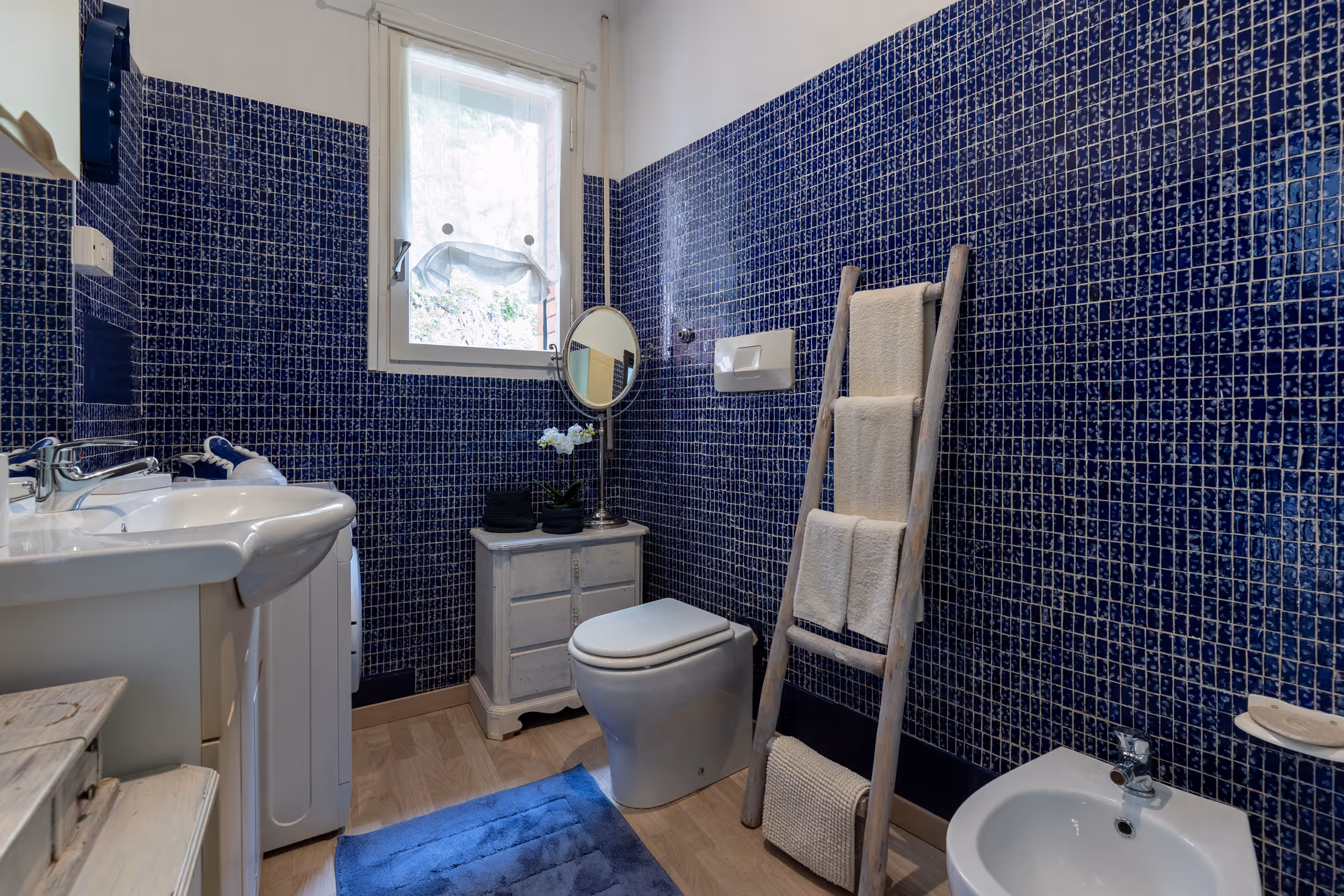 Bagno moderno con piastrelle blu scuro, lavandino bianco, toilette e scala portasciugamani.