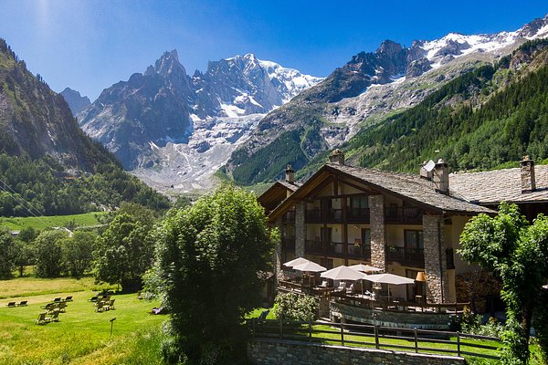 Rifugio di montagna in valle alpina con picchi innevati, prati verdi e alberi.