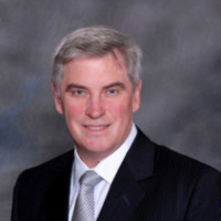Professional headshot of a middle-aged man with gray hair wearing a black suit, white shirt, and light gray tie against a gray studio background.