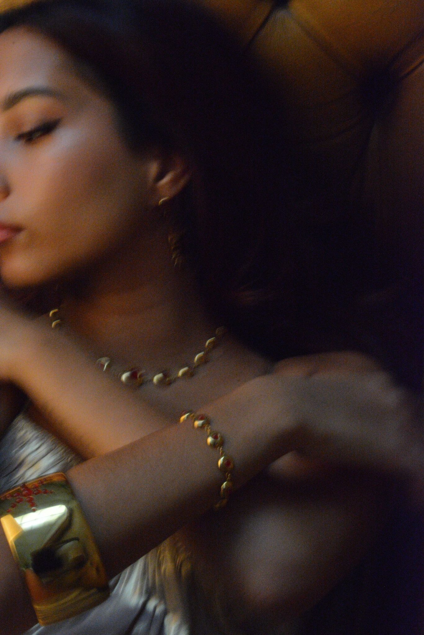 Blurred close-up of a woman wearing gold jewelry including a bracelet, necklace, and earrings.