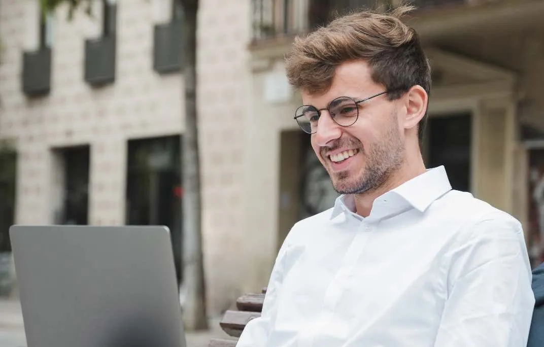 Ein junger Mann im weißen Hemd sitzt draußen in einem Kaffee und arbeitet gut gelaunt vor seinem Laptop.
