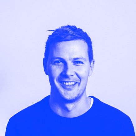 An image of a man with short side-swept hair, wearing a black jumper smiling at the camera. Blue filter on the image. 