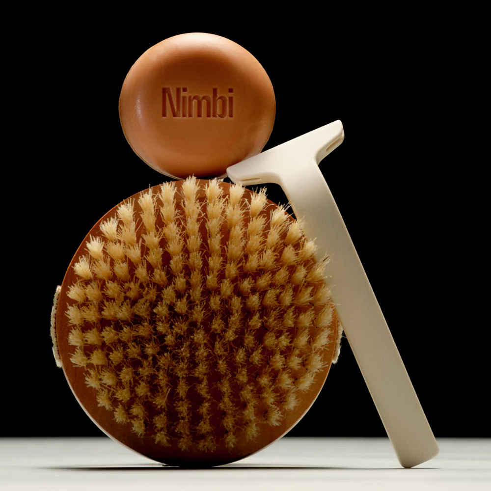 A exfoliating brush, Nimbi-branded soap and face razor balanced in a studio environment. Black background.