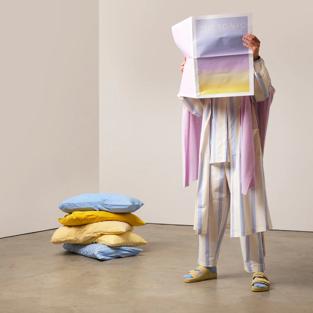 A person in pastel coloured pyjamas holding a newspaper high in front of their face. A stack of cushions in the background.