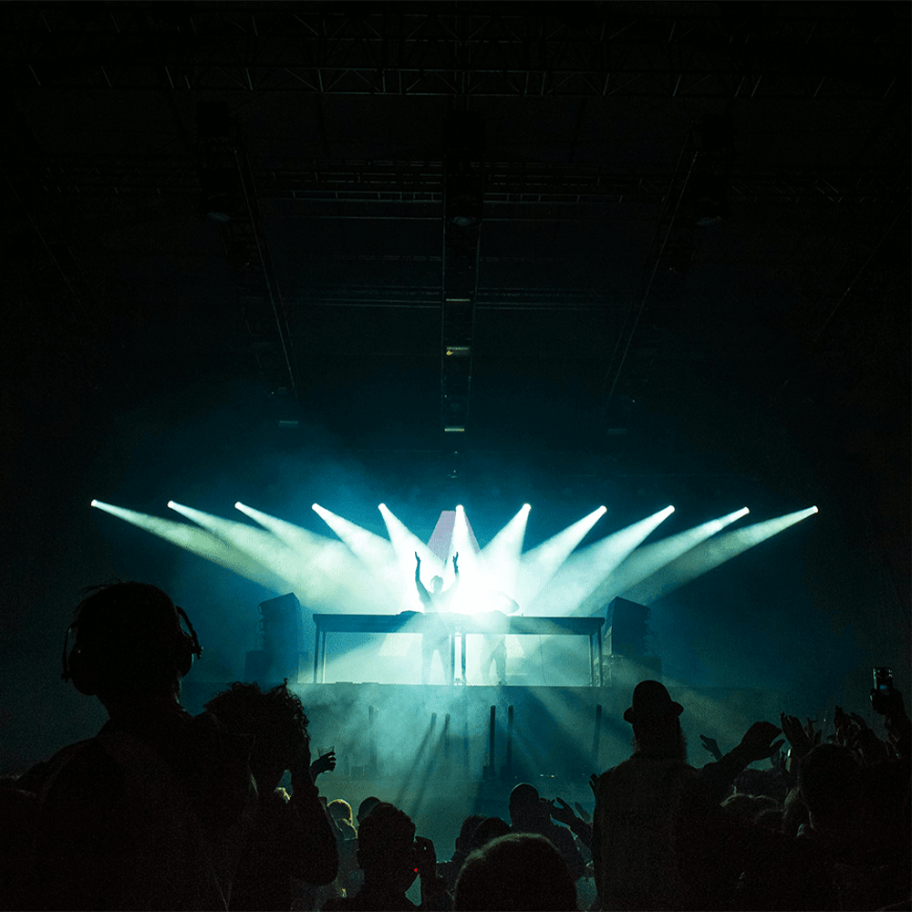 A DJ set with blue focal lighting on the stage. A crowd in the foreground dancing.