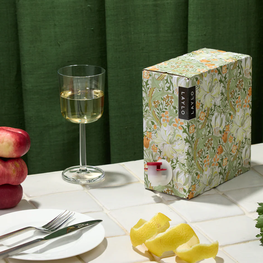 A green floral print box of wine on a dining table with a green background. Glass of white wine, cutlery and peaches in the foreground.