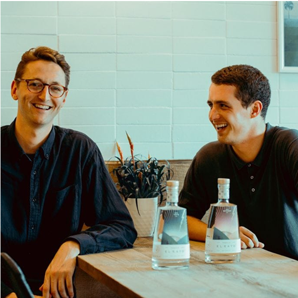 El Rayo Founders: Two laughing men leaning on a wooden table, with two bottles of tequila in the foreground. 