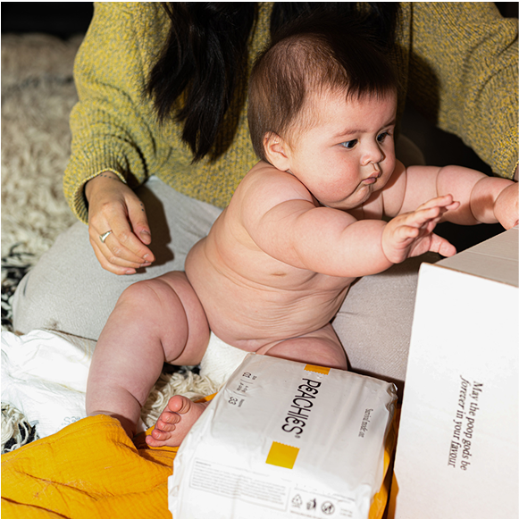 A baby in a woman's lap. Baby is curiously reaching for a package of nappies. 