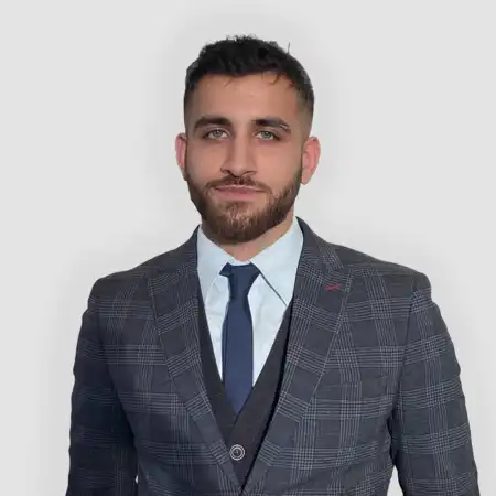 A man with short dark hair and a beard wearing a dark plaid blazer, black vest, light blue shirt, and navy tie against a plain light gray background.
