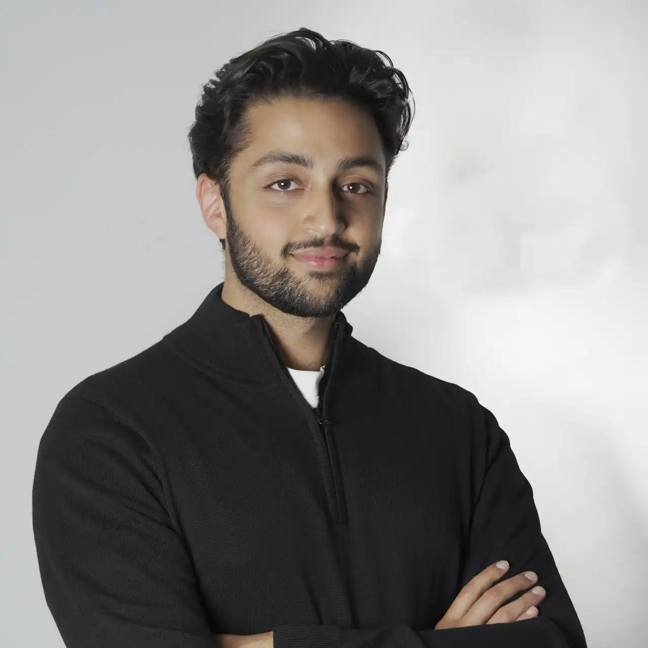 Portrait of a young man with dark hair and beard wearing a black zip-up sweater, standing with arms crossed and smiling slightly against a light gray background.