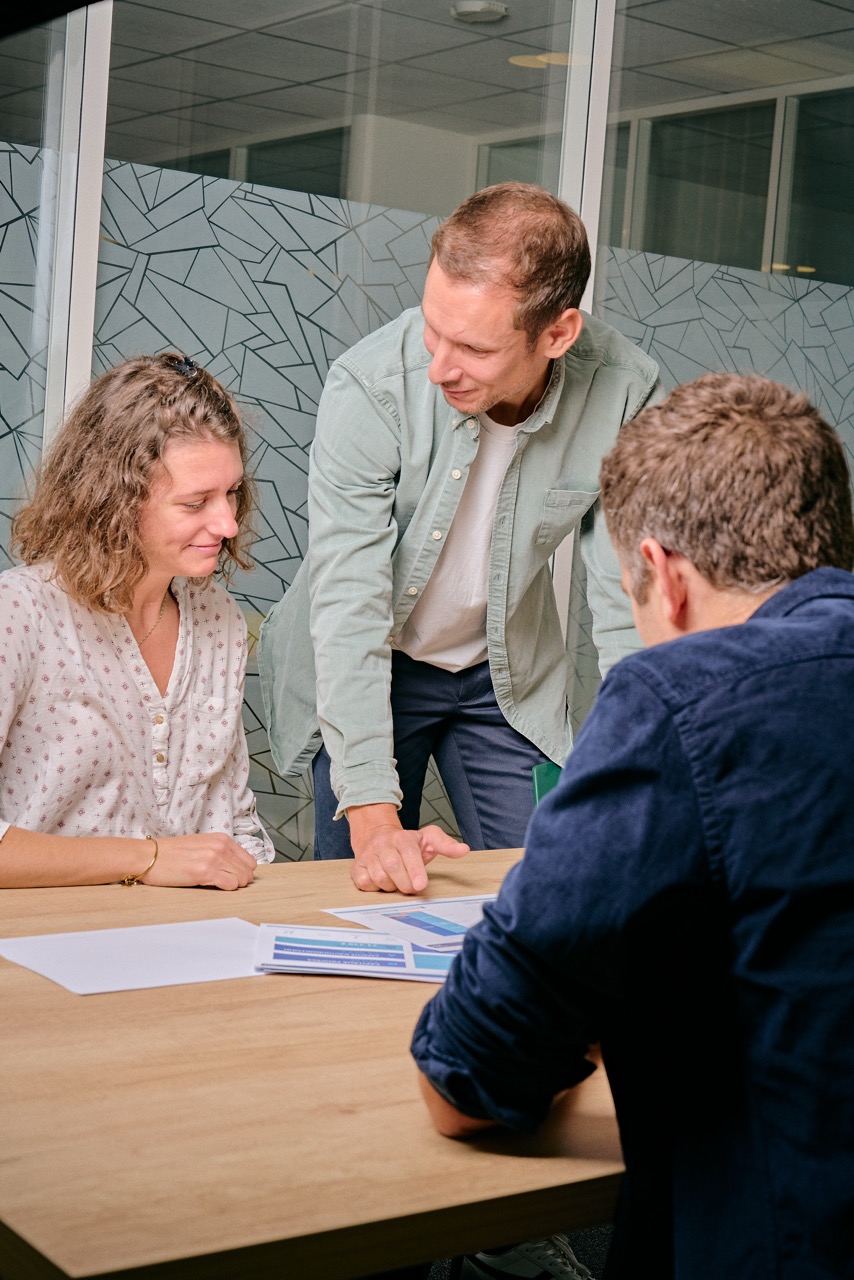 Trois collègues discutant autour d'une table avec des documents, un homme debout montrant des données sur une feuille.