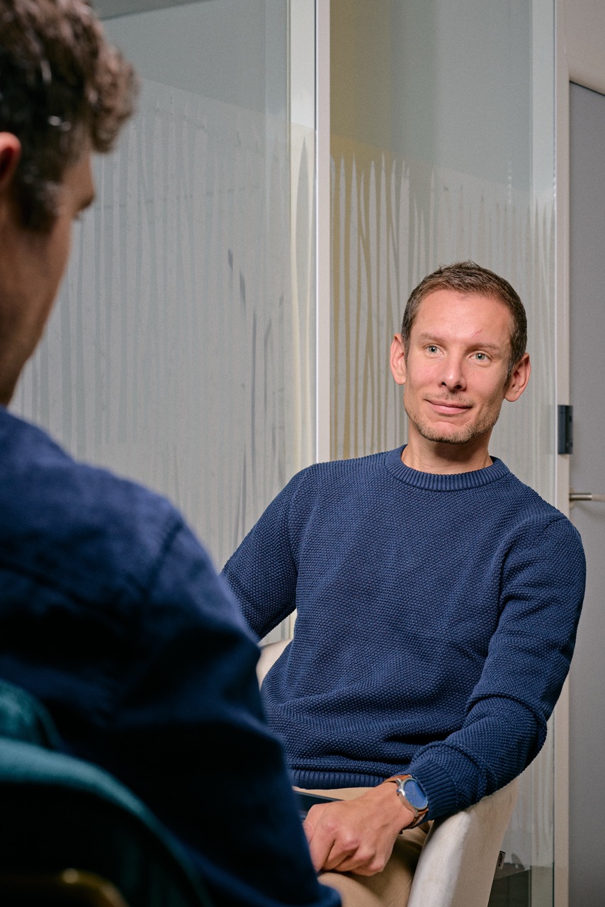 Homme assis en pull bleu avec une montre, écoutant une autre personne pendant une discussion dans un bureau moderne.