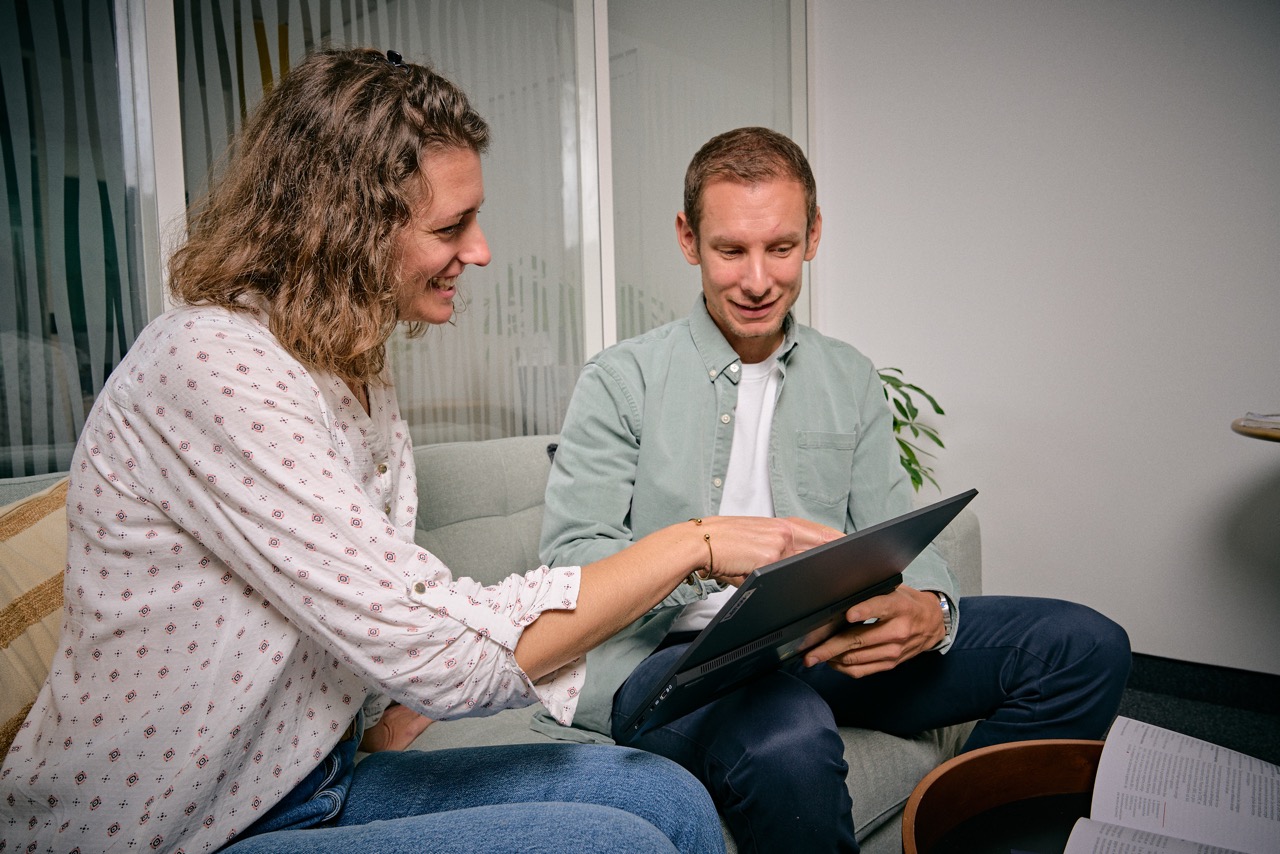 Deux personnes assises sur un canapé regardant un ordinateur portable, la femme pointe quelque chose sur l'écran en souriant.
