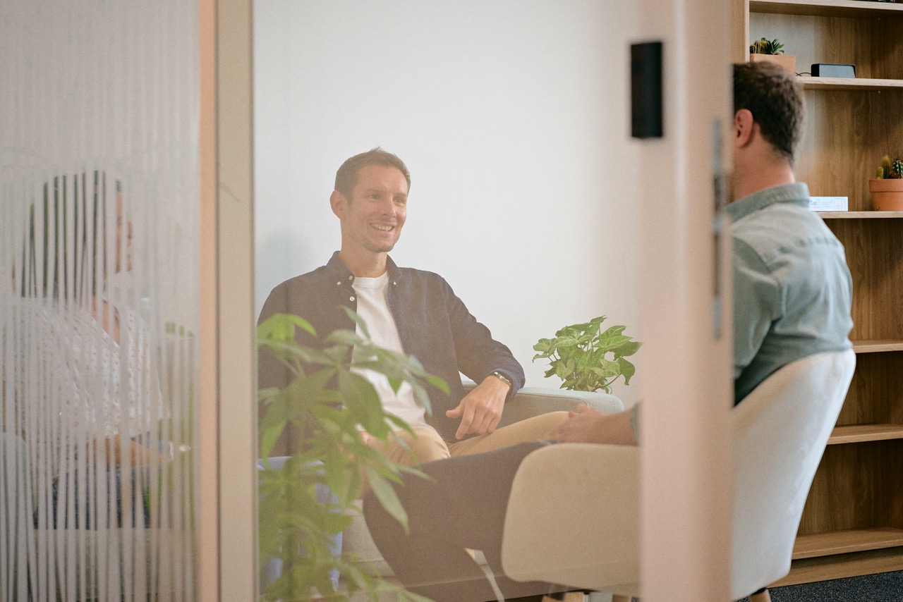 Trois hommes assis dans une salle de réunion moderne avec des plantes vertes, discutant et souriant.