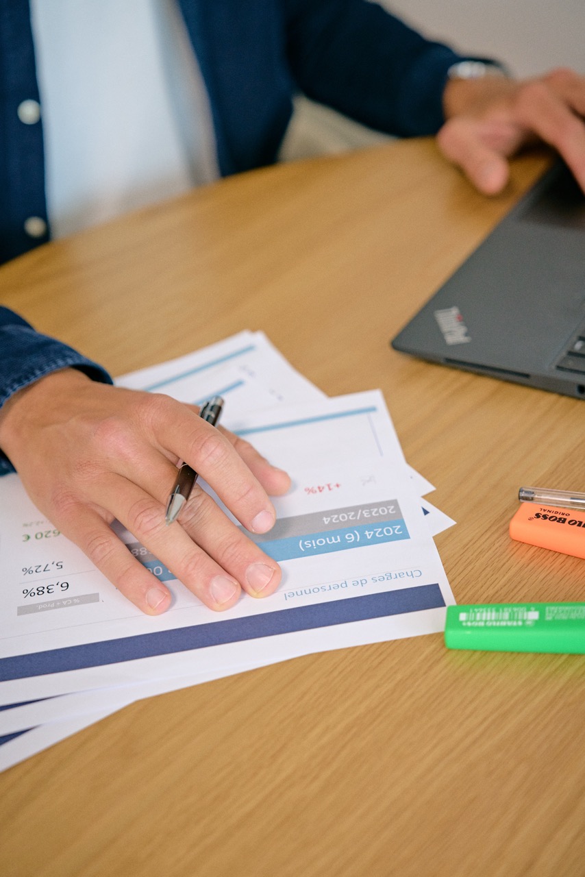 Une personne tenant un stylo au-dessus de documents avec graphiques financiers sur une table en bois à côté d'un ordinateur portable et deux surligneurs.