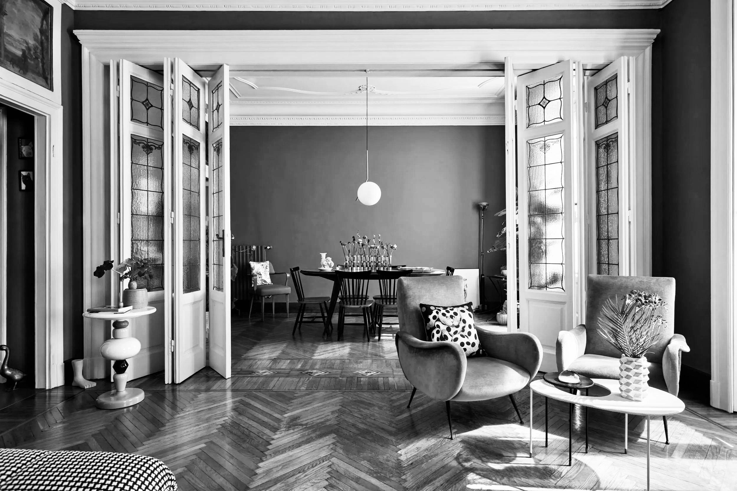 Интерьер гостиной с двумя креслами, круглым столиком с вазой и цветами, а также обеденным столом с четырьмя стульями на фоне декоративных складных дверей.