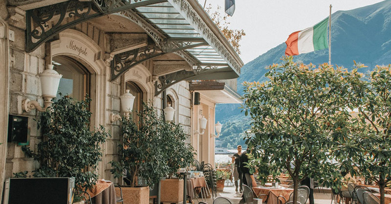 Уютная наружная терраса кафе с зелеными деревьями и итальянским флагом на фоне гор.