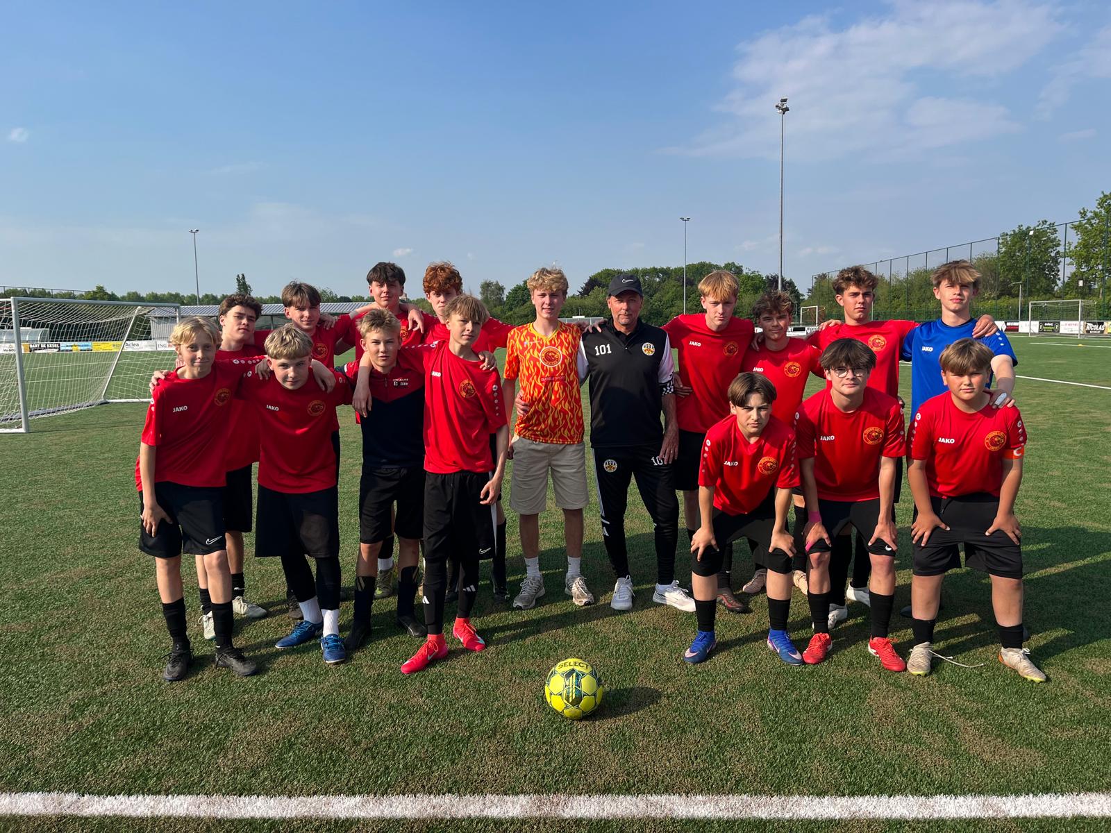 Youth soccer team of fifteen boys and one coach posing on a green soccer field in sunlight, with two goalposts visible in the background; most boys wear red jerseys and black shorts, two wear blue goalkeeper jerseys, and one has a red and yellow patterned jersey.