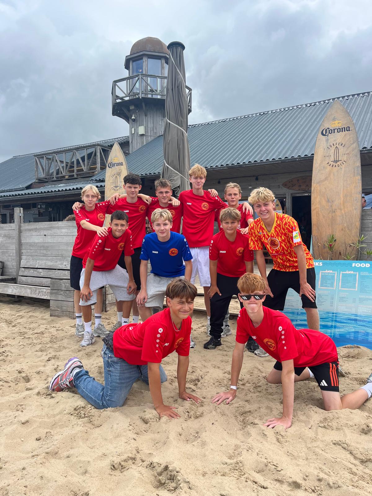 Group of eleven boys wearing mainly red sports jerseys posing on a sandy beach in front of a rustic wooden building with a small tower, surfboards with Corona branding, and a menu board.