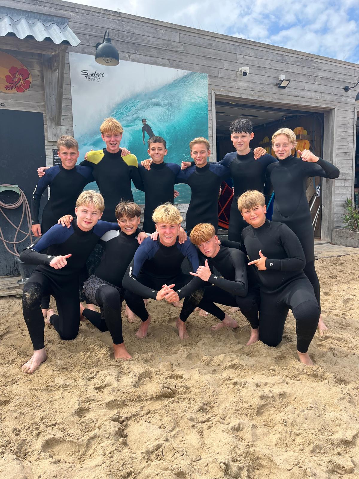 Group of eleven teenage boys wearing black wetsuits posing together on sandy beach in front of a wooden building with a large surf photo poster and some surfboards inside.